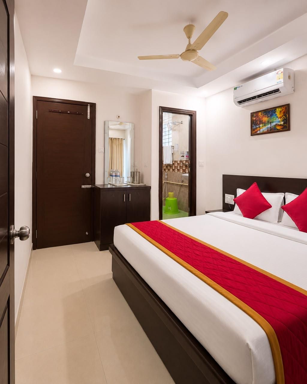 Una habitación de hotel con cama, ventilador de techo, unidad de aire acondicionado, una puerta y un baño visible a través de una puerta abierta.