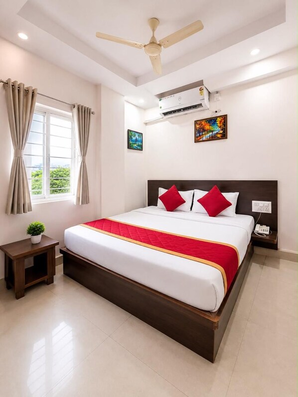 A hotel room with a bed, a nightstand, a ceiling fan, an air conditioning unit, a window with curtains, and a painting on the wall.