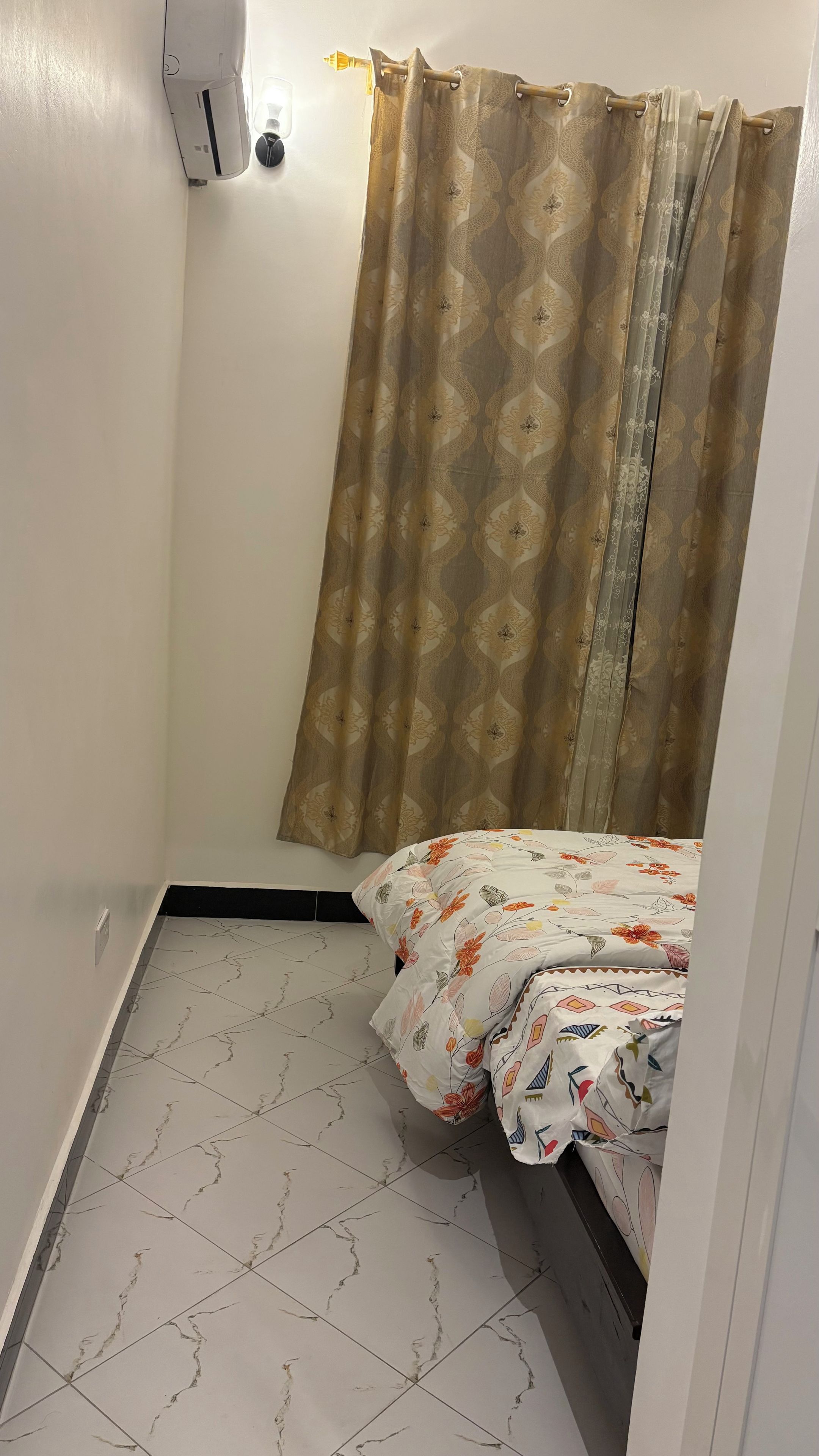 A small, single-bed room with a patterned bedspread, a curtain, and a tiled floor.
