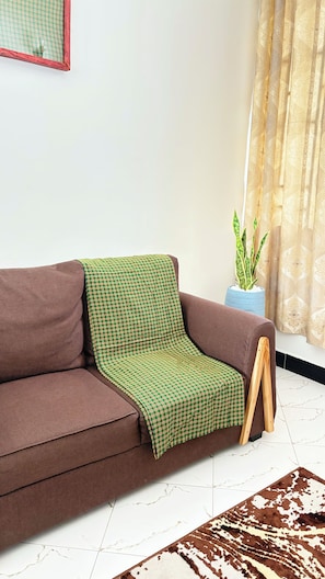 Ein braunes Sofa mit einer grün karierten Decke, eine blaue Vase mit einer Pflanze und ein gemustertes Teppich auf einem weiß gefliesten Boden.