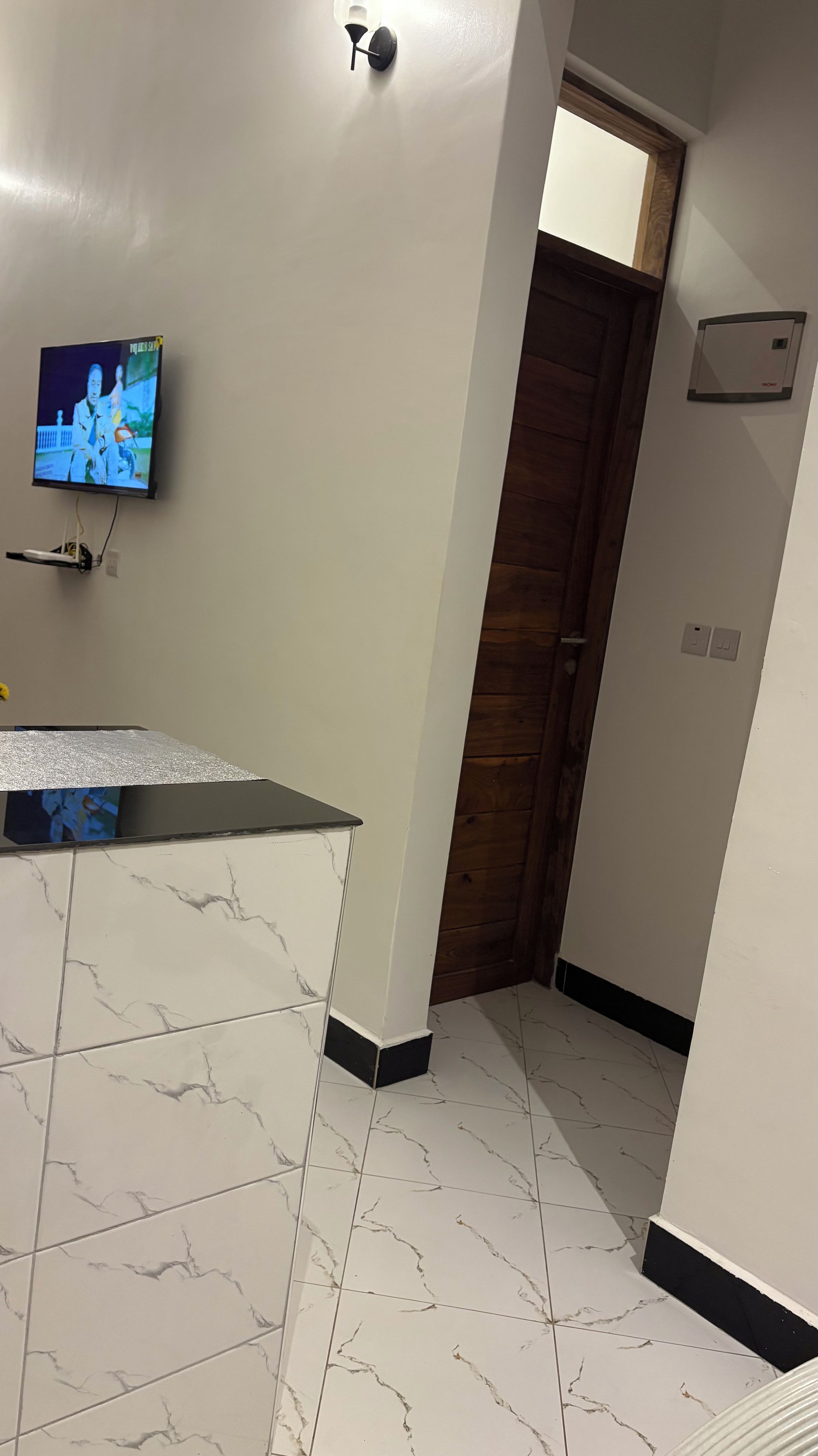 A room with a television mounted on the wall, a wooden door, and a tiled floor with a marble pattern.