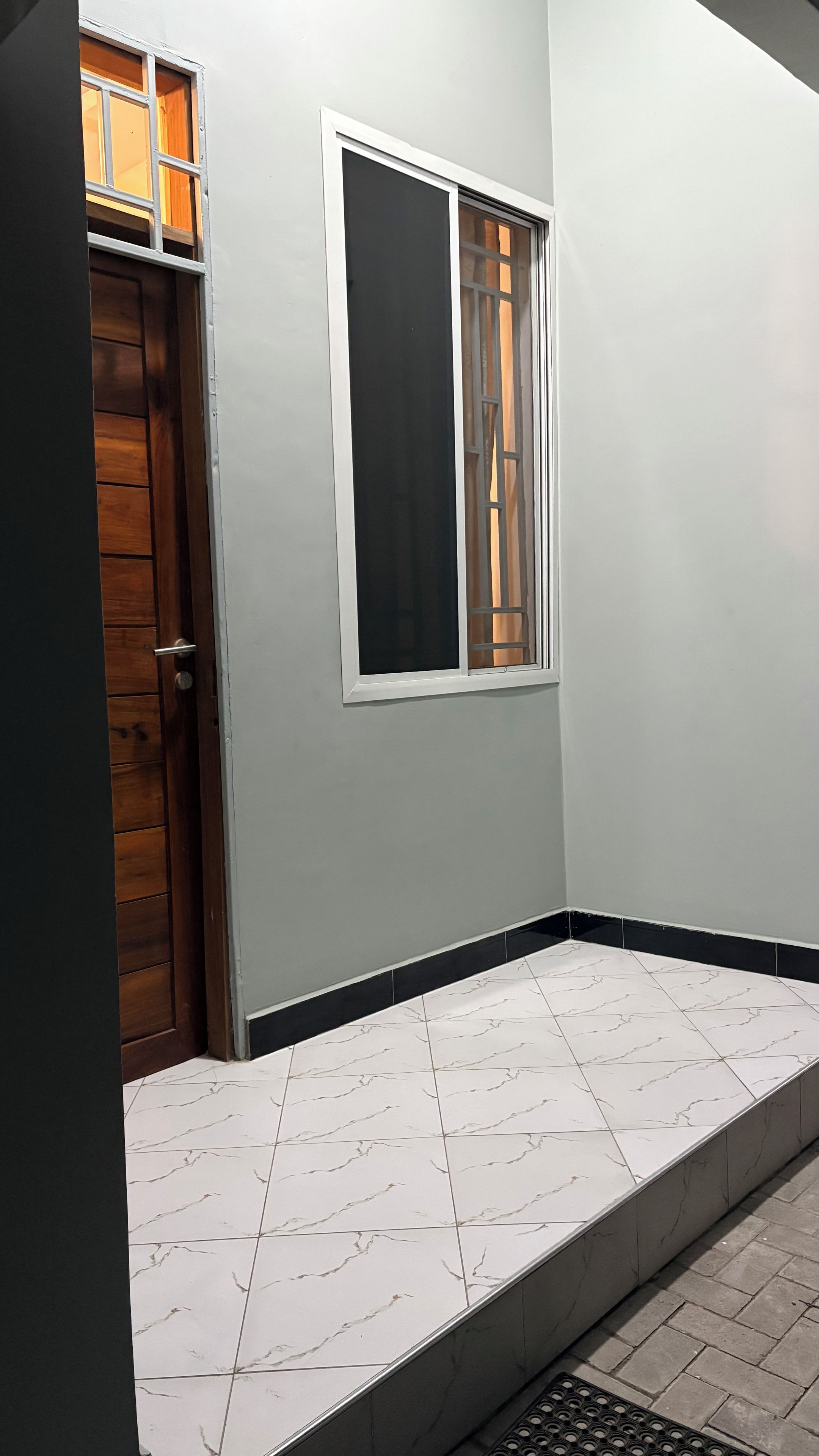 Um quarto com soalho em azulejo, porta de madeira e janela com moldura branca.