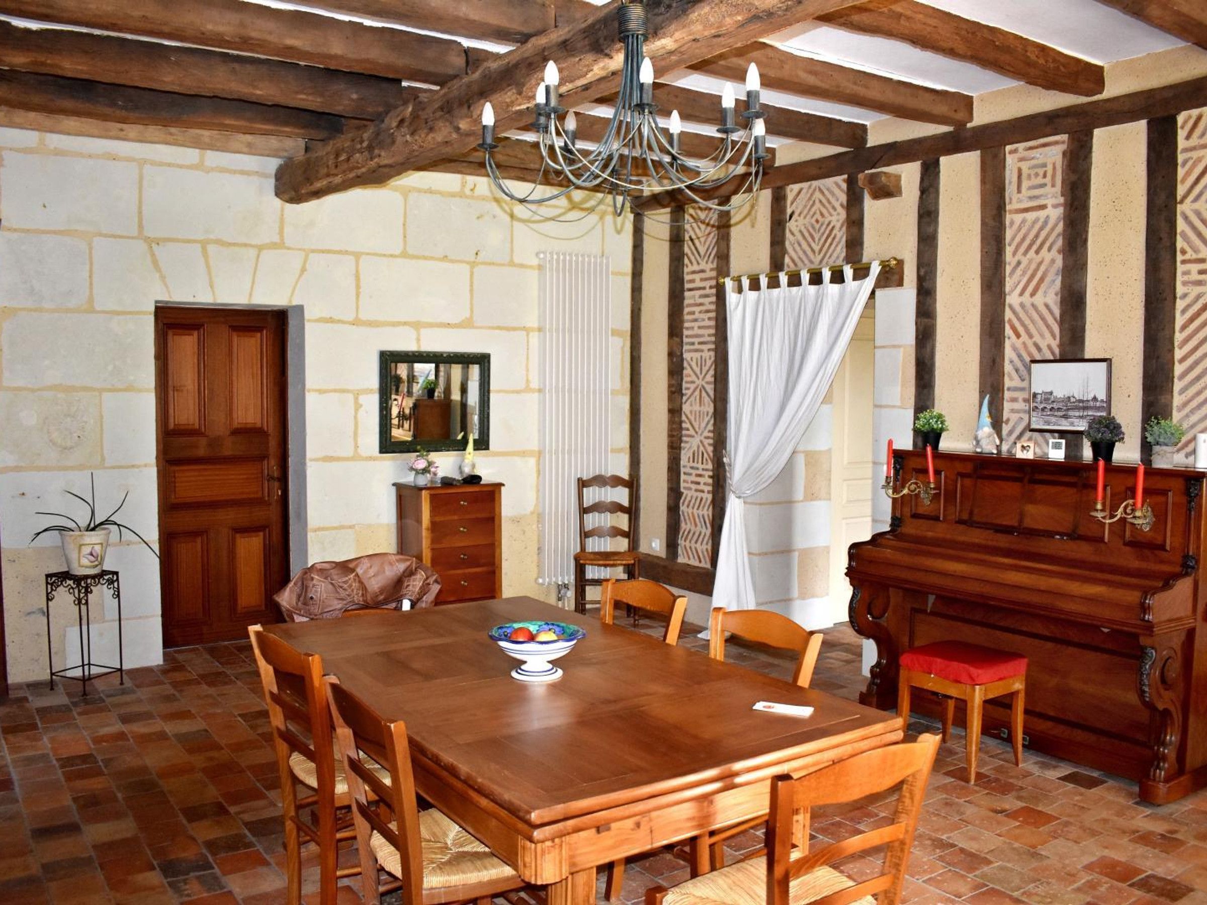 A dining area with a wooden table, chairs, a chandelier, a piano, and a stone wall.