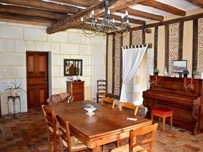 A dining area with a wooden table, chairs, a chandelier, a piano, and a stone wall. - Charming cottage in the heart of the Loir Valley (SAINT ARNOULT)