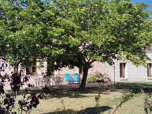 A courtyard with a large tree, outdoor seating, and a building in the background. - Charming cottage in the heart of the Loir Valley (SAINT ARNOULT)