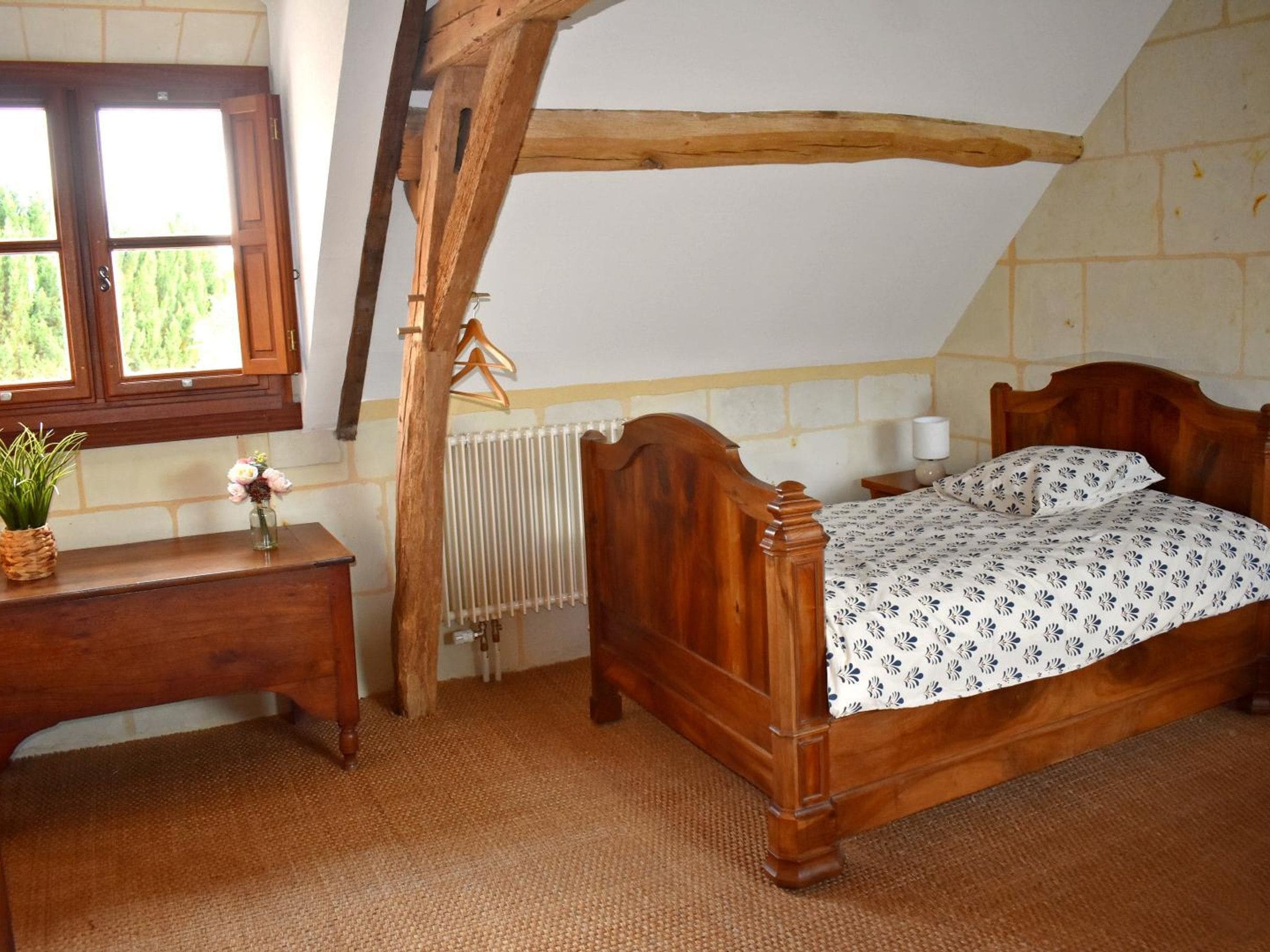 A bedroom with a wooden bed, a bedside table, a hanging coat rack, a radiator, and a window with a view of greenery.