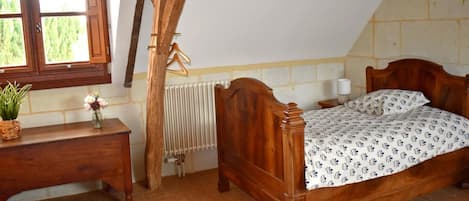 A bedroom with a wooden bed, a bedside table, a hanging coat rack, a radiator, and a window with a view of greenery.