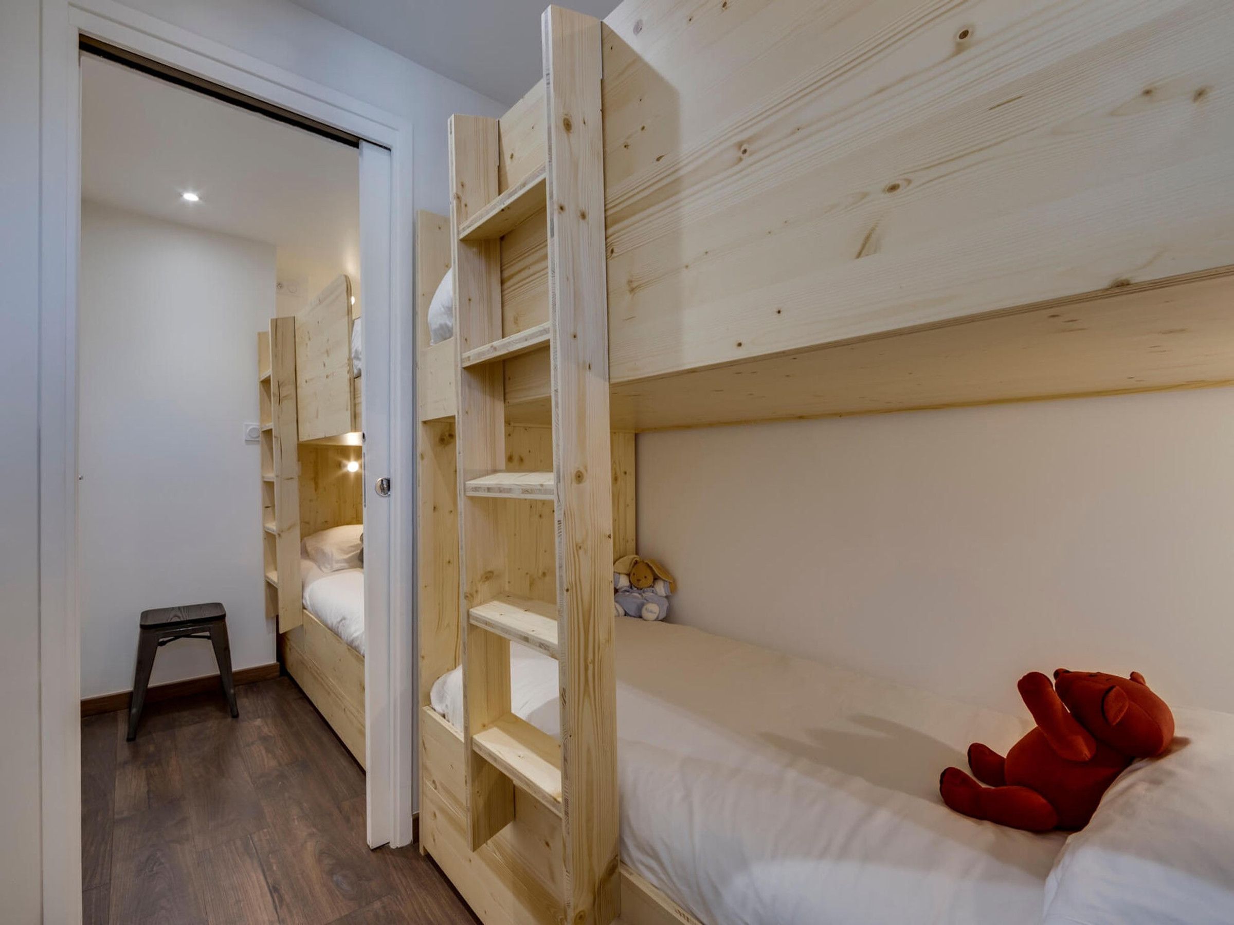 A room with bunk beds, a small stool, and a teddy bear on the bed.