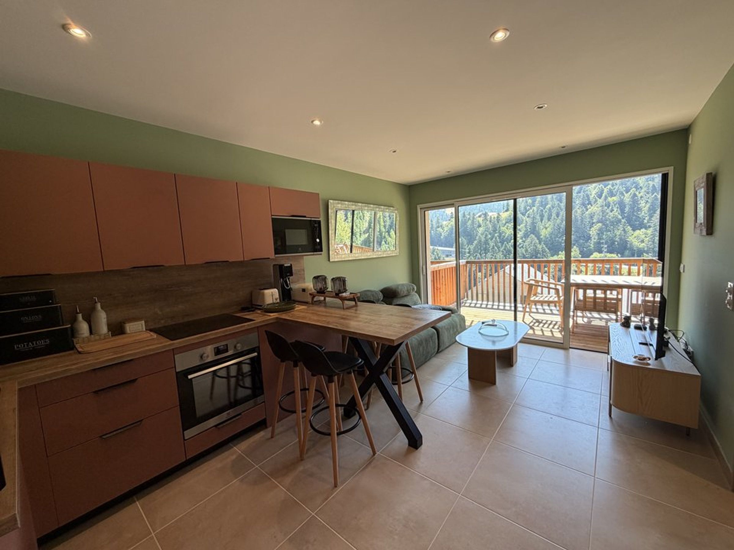 Une cuisine moderne avec un coin repas, une grande porte-fenêtre coulissante donnant sur une terrasse et une vue sur une zone boisée.
