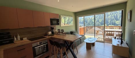 Eine moderne Küche mit Essbereich, eine große Schiebeglastür, die auf eine Terrasse führt, und Blick auf ein bewaldetes Gebiet.