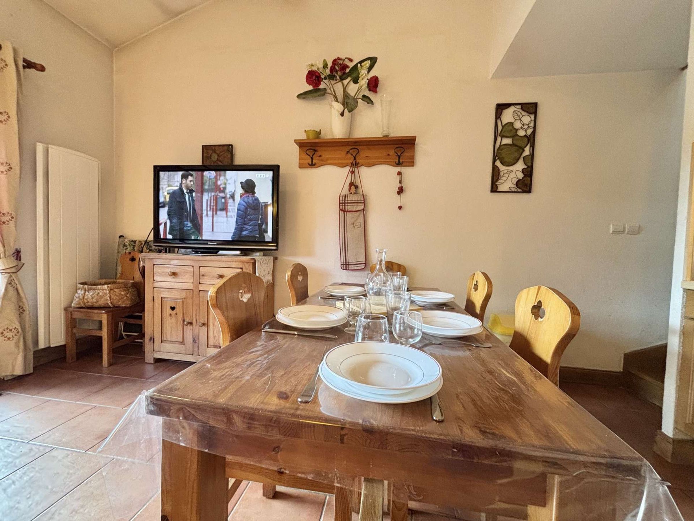 Un comedor con una mesa de madera dispuesta para cuatro, un televisor montado en la pared y un florero con flores en un estante sobre la mesa.