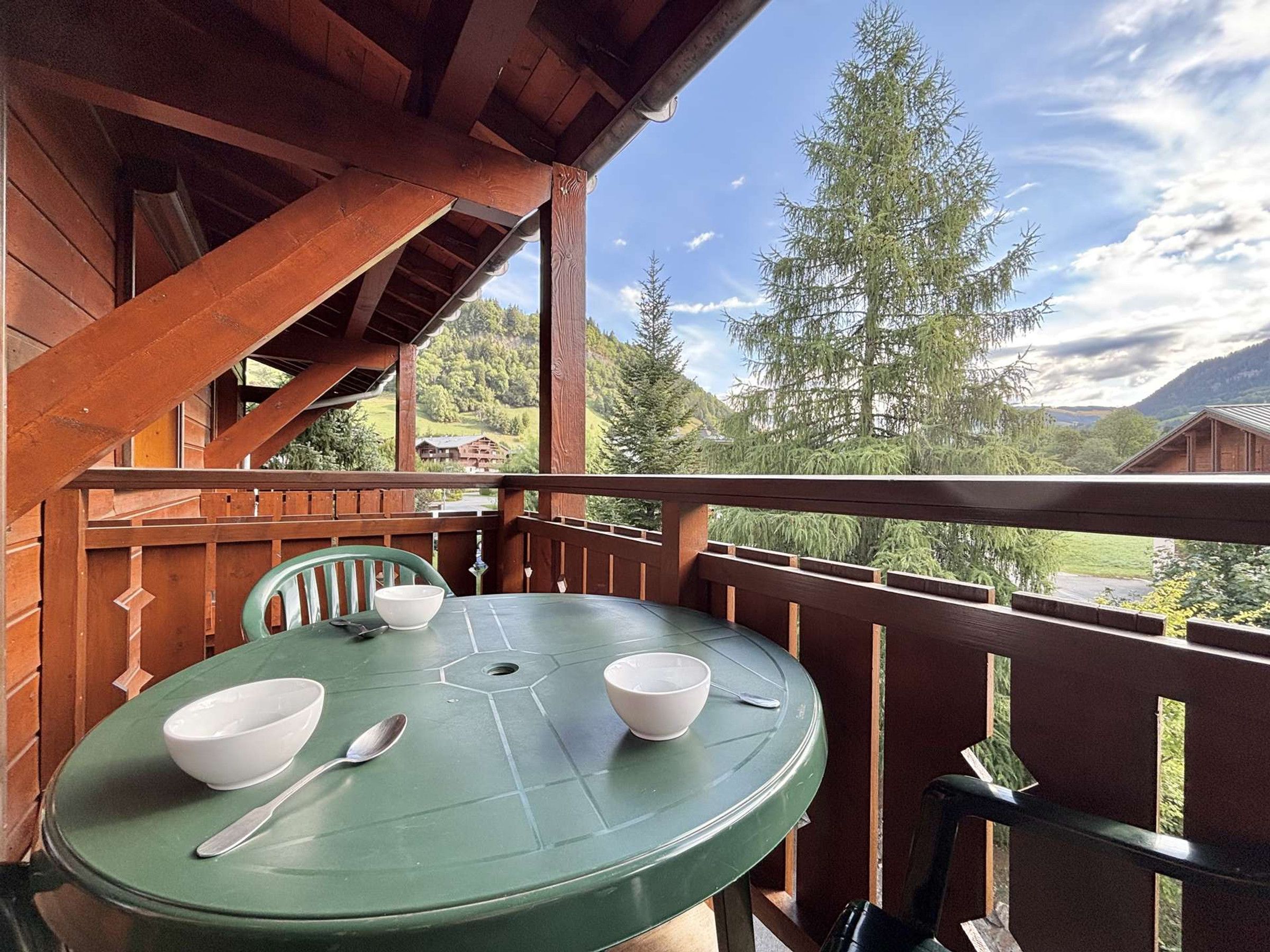 Un balcón de madera con una mesa y sillas verdes, con vistas a un paisaje montañoso.