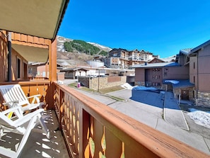 A balcony with white chairs, a stone wall, and a view of a town with buildings and a mountain. - Spacious apartment, balcony - 4 guests (Les Menuires)