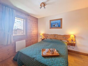 A bedroom with a wooden headboard, a bed with a green quilt, two bedside lamps, a window with curtains, and a painting on the wall. - Spacious apartment, balcony - 4 guests (Les Menuires)