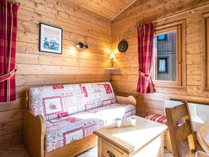 A cozy cabin interior with a wooden wall, a sofa with patterned bedding, a table with two mugs, and a framed picture on the wall. - Spacious 2-room apartment with terrace - 4 people (Les Menuires)