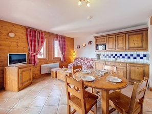 A wooden interior with a dining table set for four, a television, and a kitchen area with wooden cabinets and a tiled backsplash. - Pleasant apartment with balcony - 4 guests (Les Menuires)