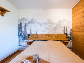 A bedroom with a wooden headboard, a bed with a patterned bedspread, a bedside table with a kettle and cup, and a wall mural of mountains. - Spacious apartment near slopes - 4 guests (Les Menuires)