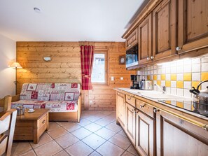 L’intérieur chaleureux d’un chalet avec un mur en bois, un canapé recouvert d’une courtepointe, une cuisine dotée de meubles en bois et une fenêtre ornée de rideaux rouges.