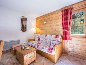 A cozy room with a wooden wall, a bed with a patterned bedspread, a small table with a kettle, and a window with curtains. - Cozy apartment near slopes - 4 guests (Les Menuires)
