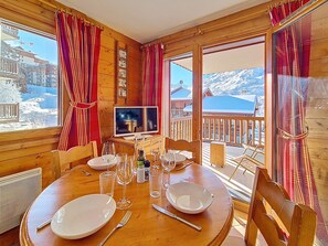 A wooden dining room with a table set for two, a television, and a view of a snowy mountain landscape. - Spacious and bright apartment, close to slopes, balcony, pool access (Les Menuires)