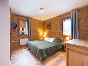 A room with wooden walls, a bed with a green blanket, a bedside table with a lamp, and a window with curtains. - Spacious apartment near slopes - 6 guests (Les Menuires)