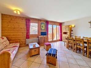 A cozy room with wooden walls, a sofa, a small table, a TV, and a dining area with wooden chairs and a table. - Spacious apartment near the slopes (Les Menuires)