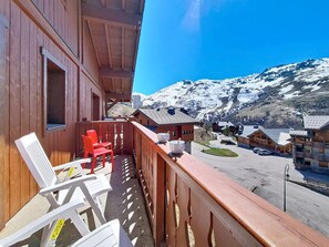 A wooden balcony with chairs and a table, overlooking a mountain landscape with snow-covered peaks and a small village. - Spacious apartment near the slopes (Les Menuires)