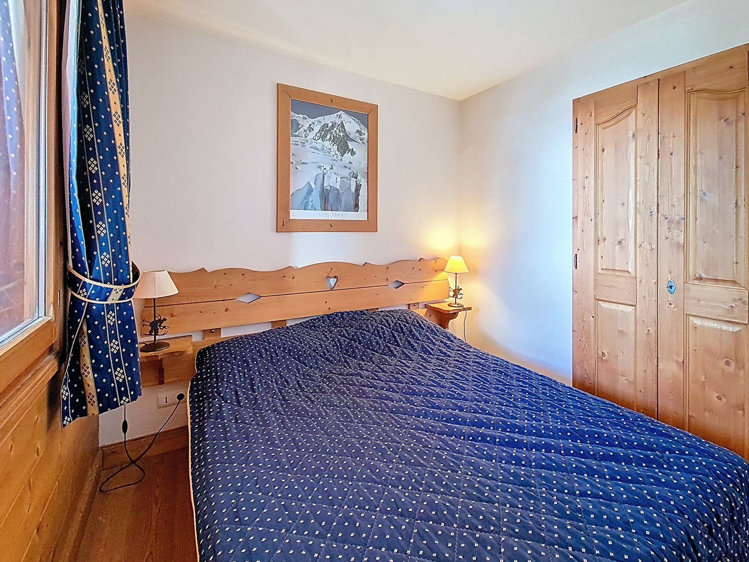 Ein Schlafzimmer mit einem hölzernen Kopfende, einem Bett mit blauer Steppdecke, einem Nachttisch mit Lampe, einem gerahmten Bild an der Wand und einem Holzschrank.