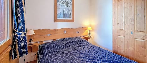 Un dormitorio con cabecera de madera, cama con colcha azul, mesita de noche con lámpara, un cuadro enmarcado en la pared y un armario de madera.