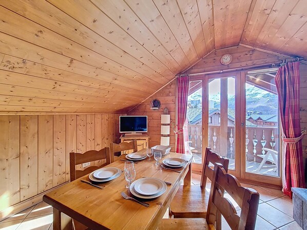 Ein Essbereich mit einem Holztisch, gedeckt für vier Personen, einem Fernseher auf einem Schrank und Blick auf eine gebirgige Landschaft durch große Fenster.
