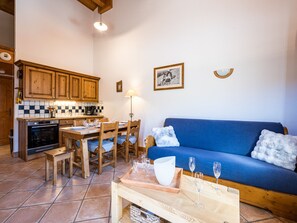 A living room with a blue sofa, wooden dining table, and chairs. There is a kitchen area with wooden cabinets and a tiled backsplash. - Spacious apartment near the slopes (Les Menuires)