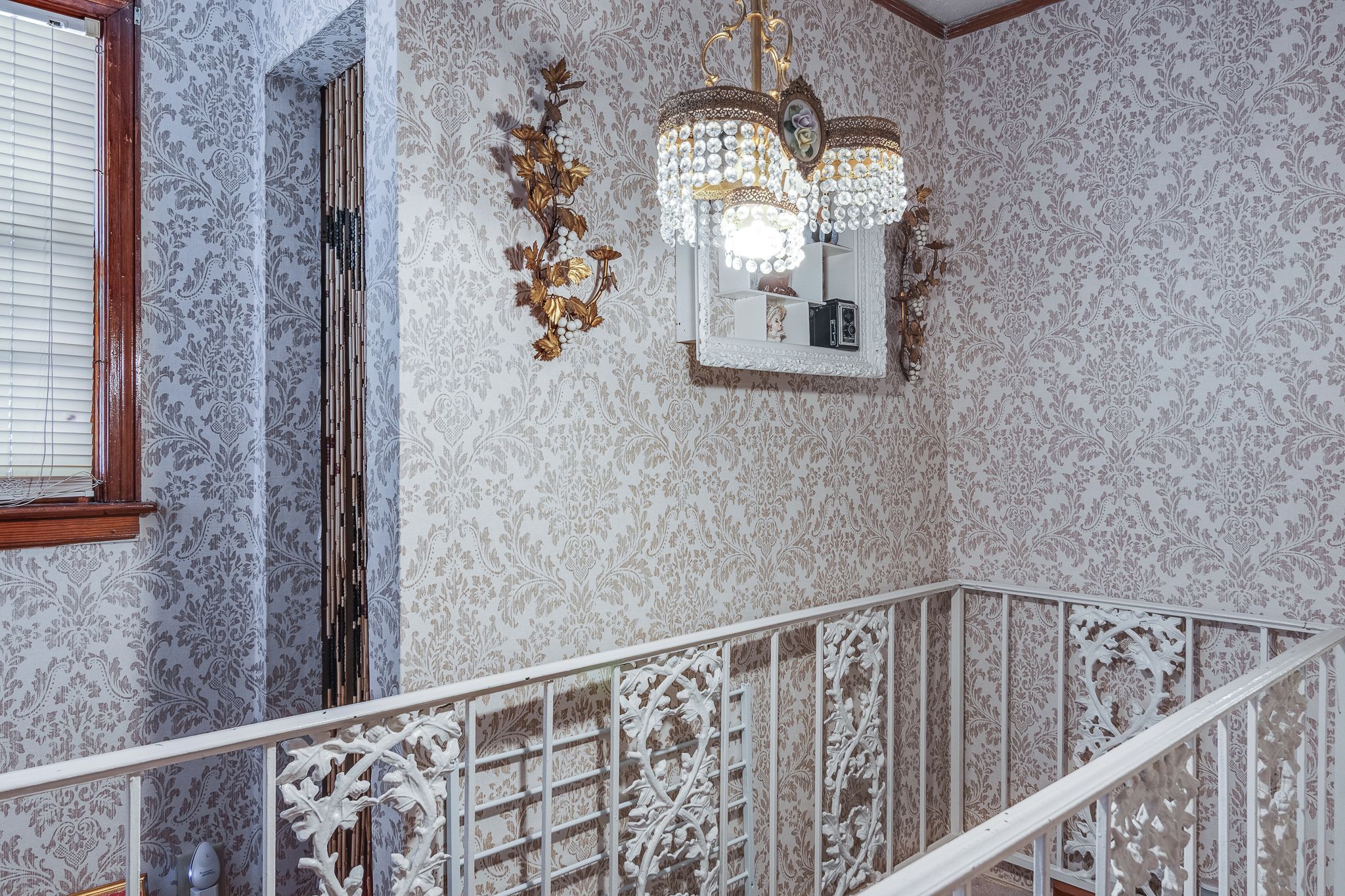 Una escalera con barandillas blancas ornamentadas, una pared con papel tapiz estampado y un espejo decorativo.