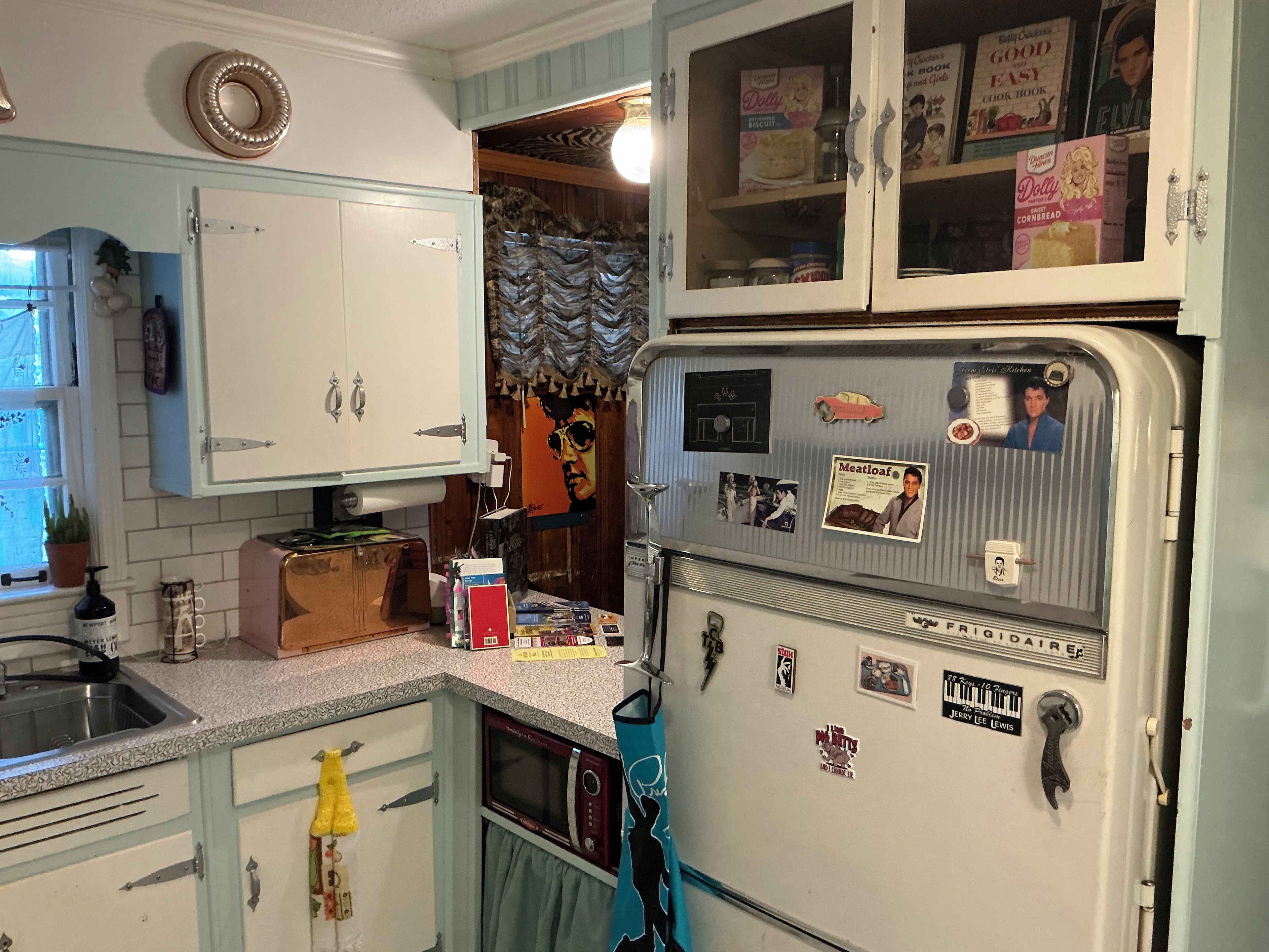 Cozinha com eletrodomésticos antigos, incluindo uma geladeira e um micro-ondas, e um armário com janela.