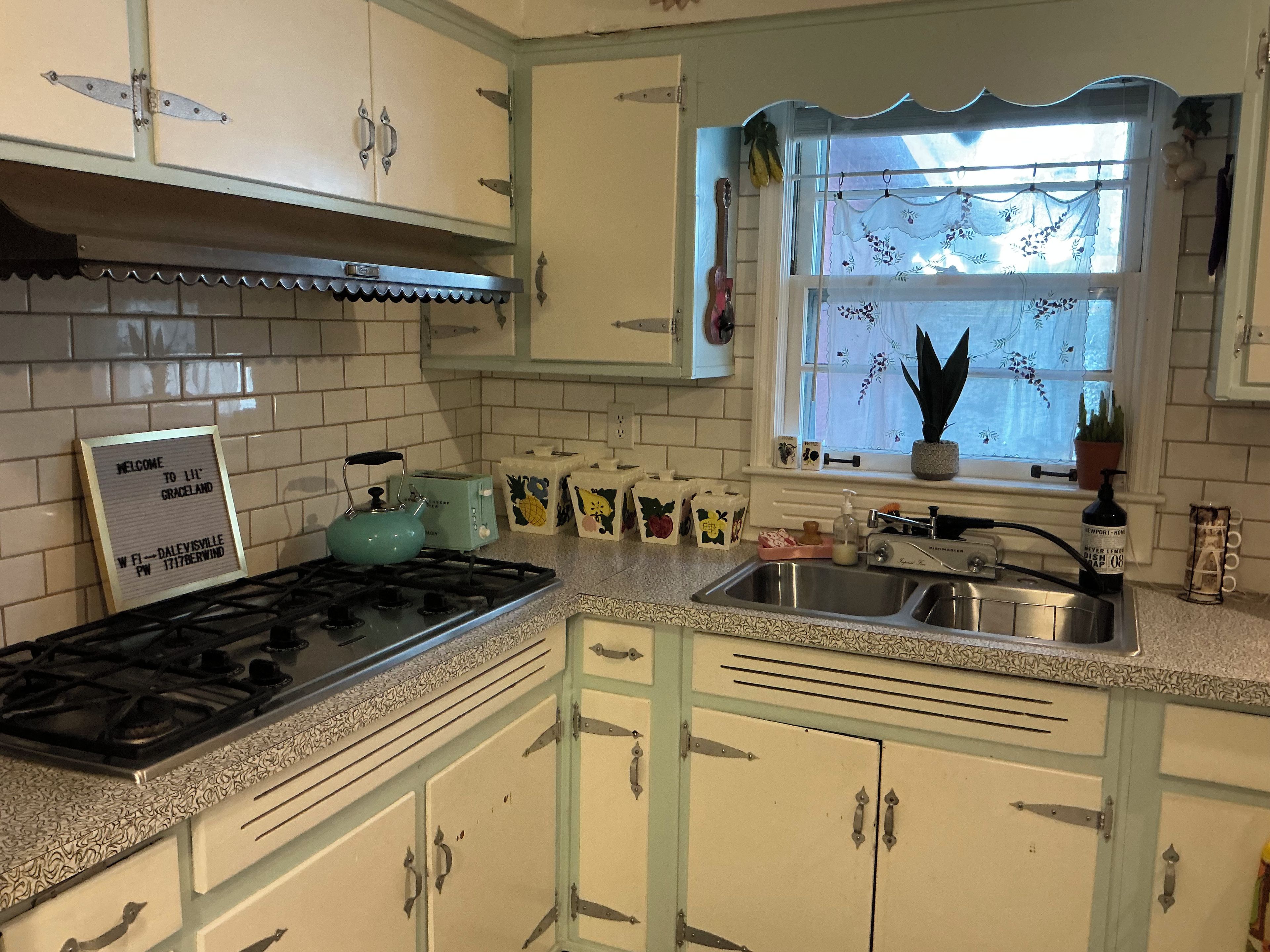 A kitchen with white cabinets, a gas stove, a window with lace curtains, and a welcome sign.