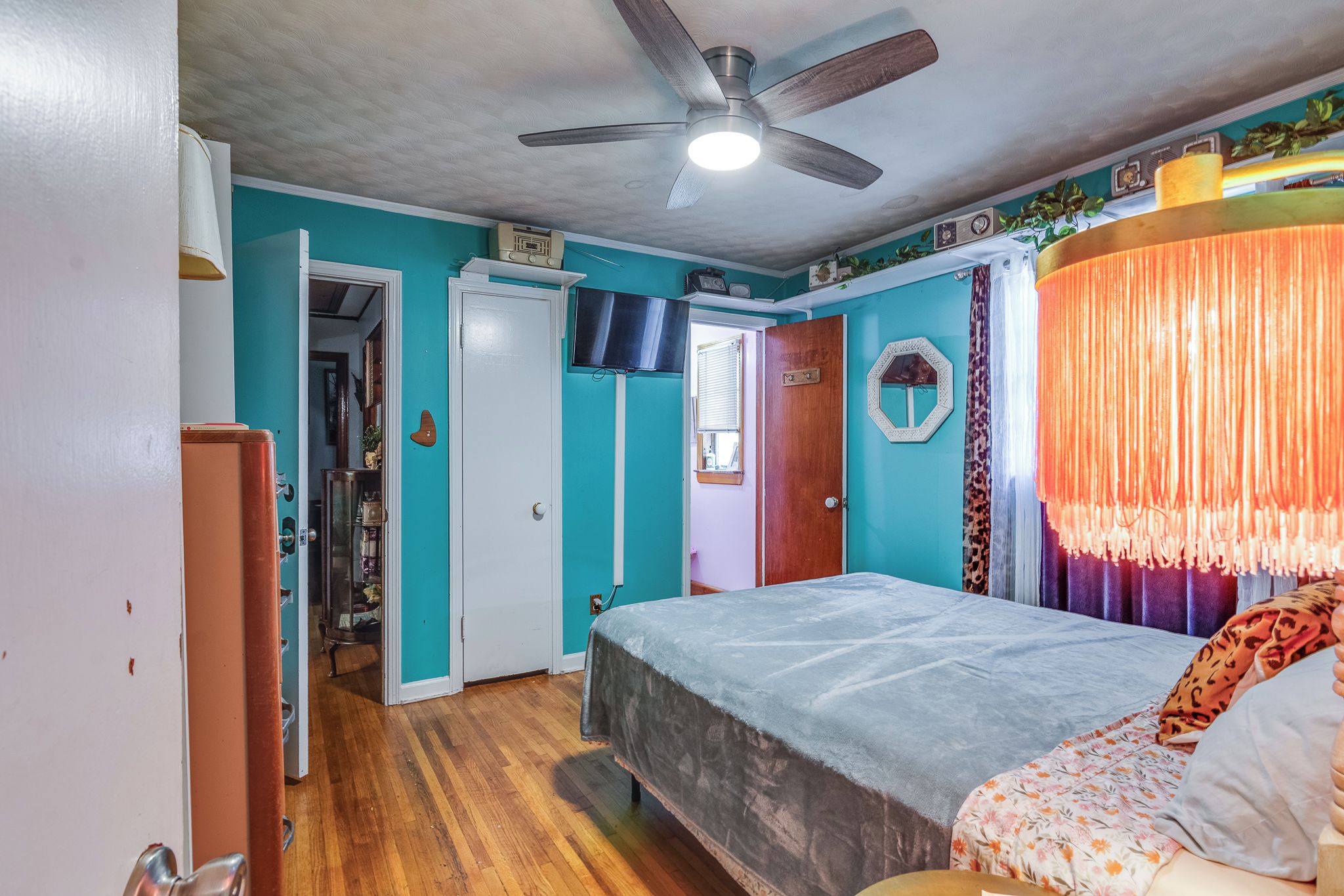 A bedroom with a bed, a ceiling fan, a television, and a door leading to another room.