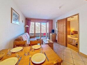 A dining area with a wooden table set for four, a sofa, and a television on a cabinet. - Bright apartment near slopes - 4 guests (Les Menuires)
