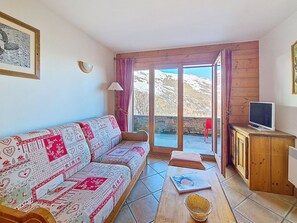A cozy living room with a patterned sofa, a television on a wooden cabinet, and a view of a mountainous landscape through a glass door. - Bright apartment near slopes - 4 guests (Les Menuires)