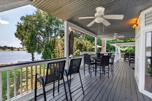Een overdekt terras met uitzicht op een meer, met een tafel, stoelen en een plafondventilator.