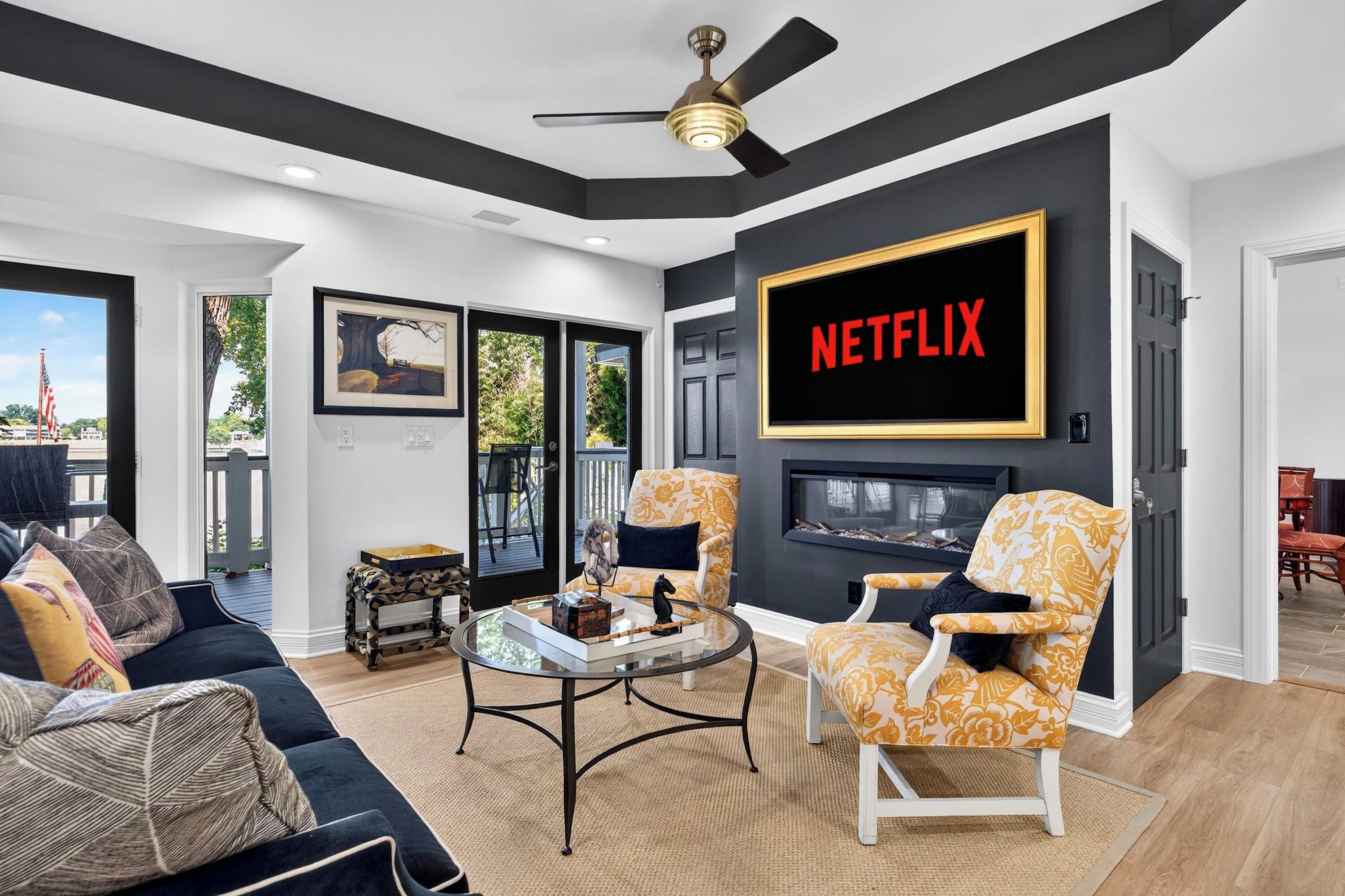 A modern living room with a fireplace, a large TV screen displaying 'NETFLIX', and a balcony with outdoor furniture.