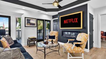 A modern living room with a fireplace, a large TV screen displaying 'NETFLIX', and a balcony with outdoor furniture.