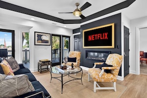 Una sala de estar moderna con chimenea, una pantalla grande mostrando 'NETFLIX' y un balcón con mobiliario exterior.