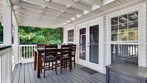 A covered outdoor deck with a table and chairs, a grill, and a view of greenery. - Premium waterfront home w/ hot tub & scenic views! (Louisville)