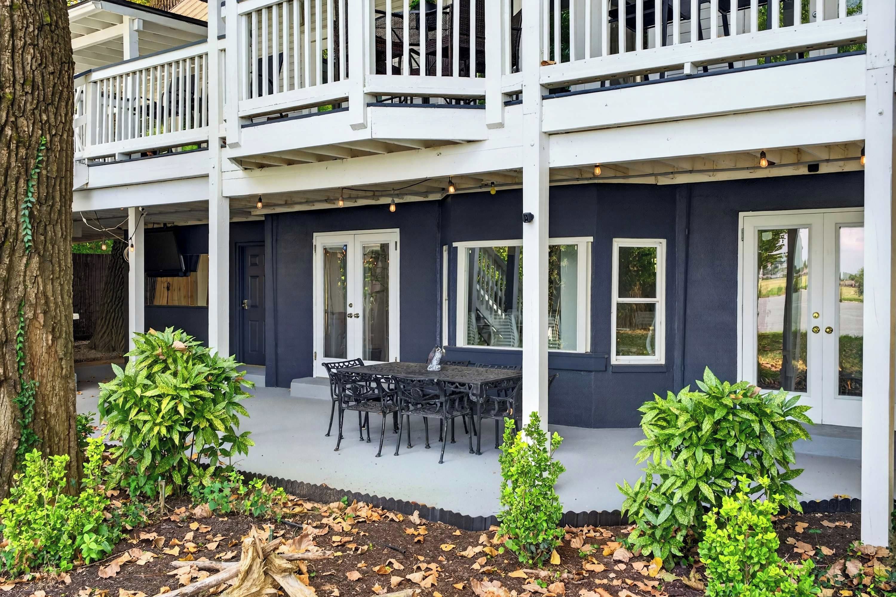 A two-story building with a balcony, a dining area with a table and chairs, and a garden with plants and fallen leaves.