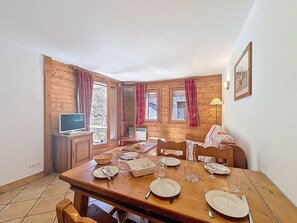 A dining area with a wooden table set for four, a television on a wooden cabinet, and a window with curtains. - Spacious apartment with balcony - 6 guests (Les Menuires)