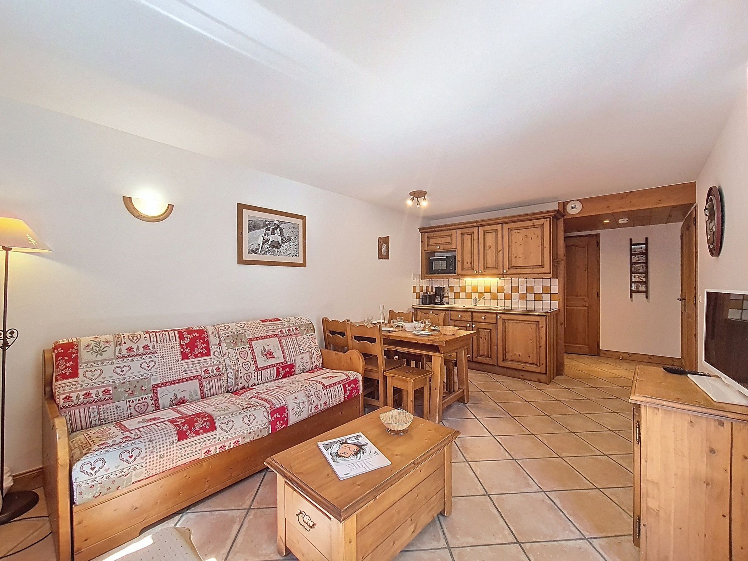 A cozy living room with a patterned sofa, wooden dining table, and a small wooden chest. A kitchen with wooden cabinets and a tiled backsplash is visible in the background.