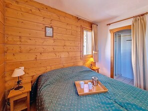 A wooden-paneled room with a bed, a nightstand, a window with curtains, and a door. - Bright apartment near the slopes - 4 guests (Les Menuires)