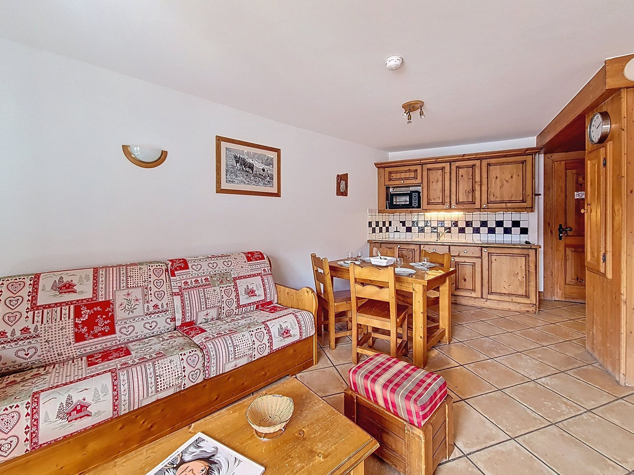 Un espacio de sala acogedor con un sofá estampado, una mesa de comedor de madera y sillas. Un pequeño banco de madera con cojín se encuentra frente al sofá. La zona de la cocina presenta armarios de madera y un salpicadero azulejado.