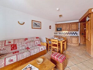 A cozy living area with a patterned sofa, wooden dining table, and chairs. A small wooden bench with a cushion is placed in front of the sofa. The kitchen area features wooden cabinets and a tiled backsplash. - Spacious apartment with balcony - 6 guests (Les Menuires)