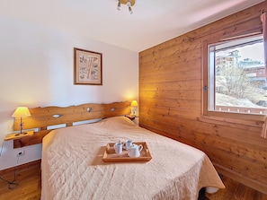 A bedroom with a wooden headboard, a bed with a beige quilt, a tray with cups and a pitcher, a wall-mounted picture, and a window with a view of buildings. - Spacious apartment with balcony - 6 guests (Les Menuires)