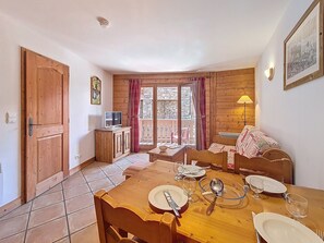 A dining area with a wooden table set for four, a television on a cabinet, and a balcony with red curtains. - Spacious apartment near the slopes - guests (Les Menuires)